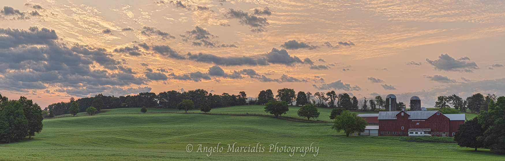 A tranquil Hudson Valley farm landscape at sunrise, with rolling green hills, scattered trees, and a red barn marked “Brookview Farm” glowing under warm orange and pink sky light.