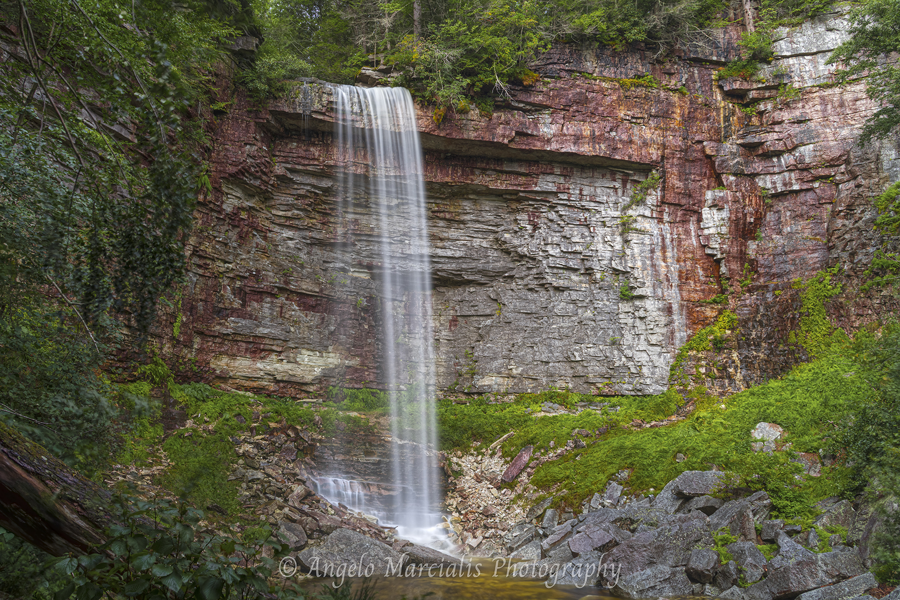 A tall, narrow waterfall descends in a smooth, long‑exposure veil over stratified red‑brown and gray rock, collecting in a small pool surrounded by moss, foliage, and dense forest greenery.