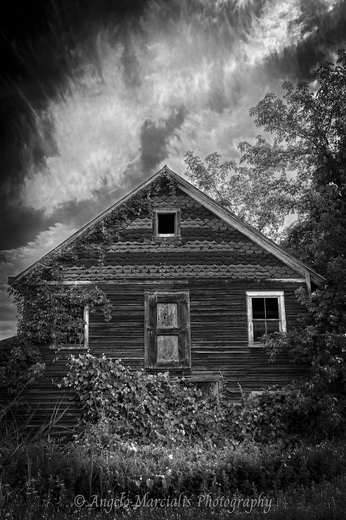 Abandoned wooden house with weathered siding, broken windows, and overgrown vines, set beneath a dramatic sky of swirling clouds and framed by surrounding trees.