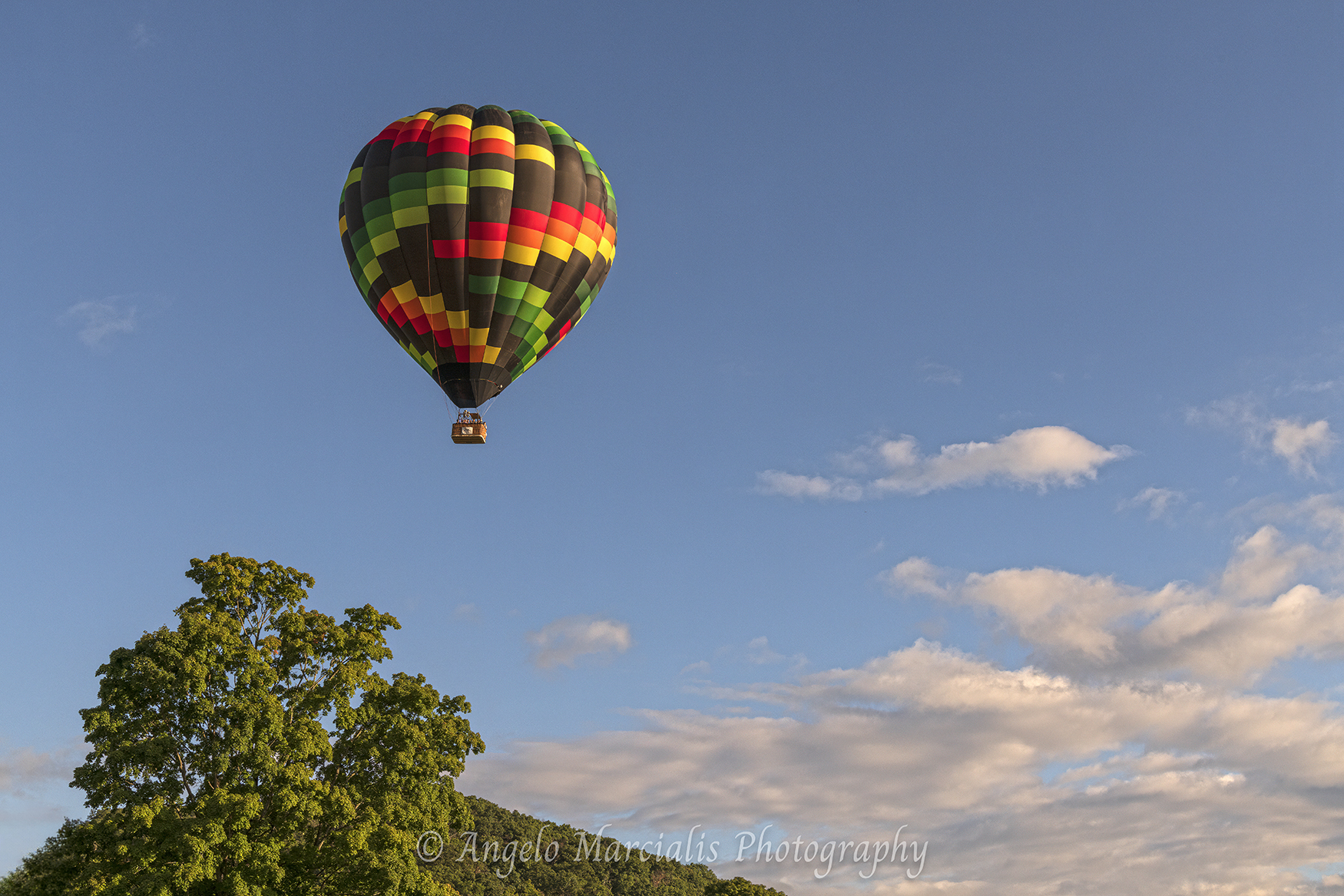 Multicolored hot air balloon soaring over the Hudson Valley, New York, above lush green trees and rolling hills beneath a bright blue sky with dramatic clouds, showcasing scenic landscape photography and aerial travel.