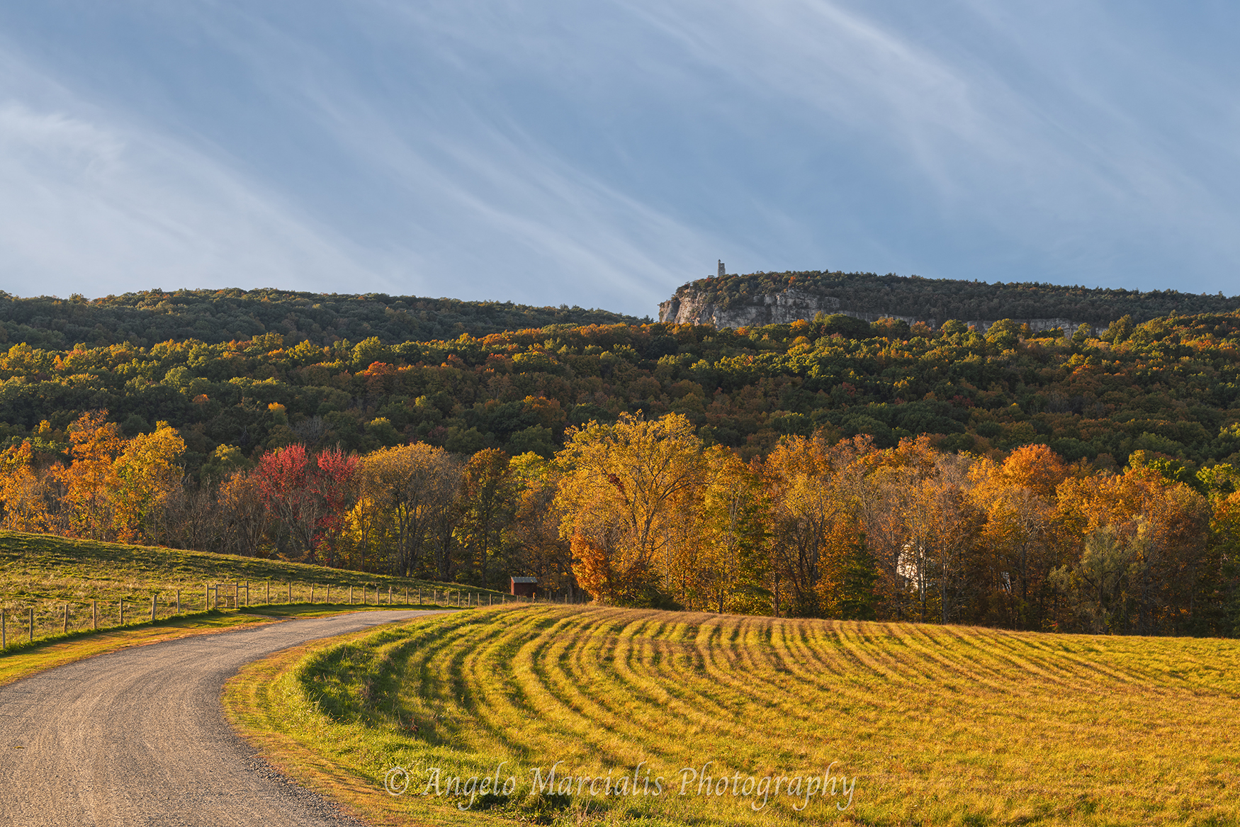 Scenic fall landscape in the Hudson Valley featuring a winding rural road, golden fields, vibrant autumn trees, and the Mohonk Mountain House tower on the Shawangunk Ridge.